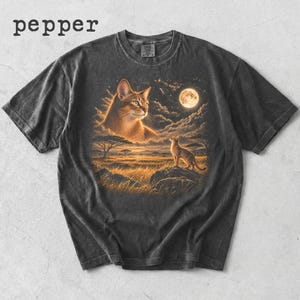 May include: A charcoal gray t-shirt with the text pepper printed in the top left corner. The center graphic features a large cat head in the clouds above a smaller cat standing on a rock in a grassy savanna landscape under a full moon.