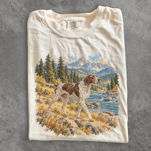 Op de afbeelding: Een crèmekleurig t-shirt met een grafische print van een bruin met wit gevlekte jachthond die op een rotsachtige rivieroever staat. De achtergrond toont een landschap met dennenbomen, een stromende rivier en besneeuwde bergen. Het label toont Comfort Colors.
