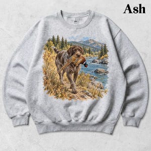 May include: An ash gray crewneck sweatshirt featuring a graphic print of a brown hunting dog carrying a pheasant in its mouth. The scene depicts the dog walking along a riverbank with pine trees, tall grass, and mountains in the background.