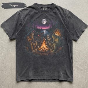 Può includere: Una t-shirt a maniche corte color grigio antracite con una stampa grafica di Bigfoot, un uomo falena, un alieno e una creatura della palude seduti attorno a un falò di notte. La scena include una luna piena, una tenda e la parola Pepper.