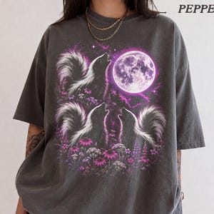 Puede incluir: Una camiseta gris carbón con un estampado gráfico de tres mofetas aullando a una luna grande y brillante de color púrpura. El diseño incluye flores y follaje púrpura alrededor de las mofetas. La palabra PEPPER está impresa en la esquina superior derecha.