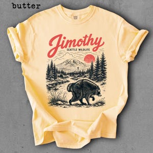May include: A butter yellow t-shirt featuring a graphic print with the text Jimothy SEATTLE WILDLIFE. The design depicts a black bear walking through a forest landscape with a mountain, a red sun, and clouds in a vintage-inspired illustration style.