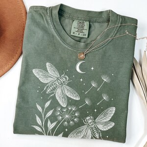 Könnte beinhalten: Ein olivgrünes Kurzarm-T-Shirt mit einem weißen Grafikdruck von zwei Zikaden, Löwenzahnsamen, einer Mondsichel und Sternen. Das Comfort Colors-Etikett am Halsausschnitt gibt die Größe S und das Material 100% Baumwolle an.