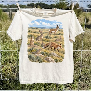 Könnte beinhalten: Ein cremefarbenes T-Shirt mit einem detaillierten Grafikdruck einer nordamerikanischen Prärielandschaft. Die Illustration zeigt einen Rotfuchs, eine Gabelbock-Antilope, einen Hasen und zwei Erdhörnchen inmitten gelber Wildblumen und Gräser.