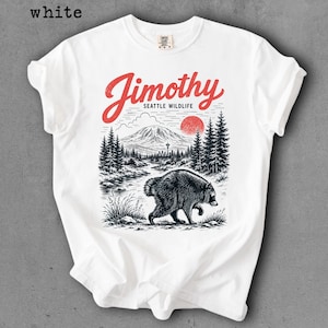 May include: A white t-shirt featuring a graphic print of a raccoon walking through a forest landscape with a mountain, a red sun, and the text Jimothy SEATTLE WILDLIFE in red script and black block lettering centered on the chest area.