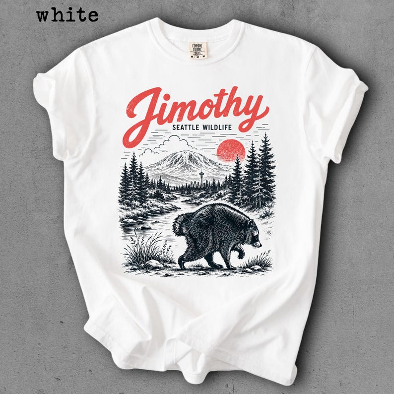 May include: A white t-shirt featuring a graphic print of a raccoon walking through a forest landscape with a mountain, a red sun, and the text Jimothy SEATTLE WILDLIFE in red script and black block lettering centered on the chest area.