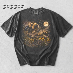 以下が含まれることがあります： 左上に「pepper」という文字がプリントされたチャコールグレーの半袖Tシャツ。中央のグラフィックには、夜の満月の下、森の風景の中で花にとまる小さなハチの上を飛ぶ大きなマルハナバチが描かれています。