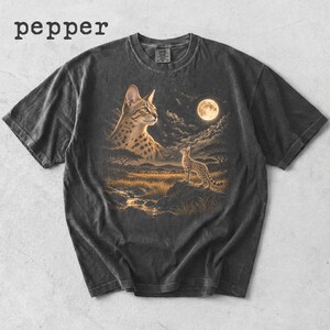 Könnte beinhalten: Ein anthrazitfarbenes Kurzarm-T-Shirt mit dem Text pepper oben links. Die Grafik zeigt eine große gefleckte Katze, die zu einer kleineren Katze auf einem Felsen in einer Savannenlandschaft unter einem Vollmond und bewölktem Nachthimmel blickt.