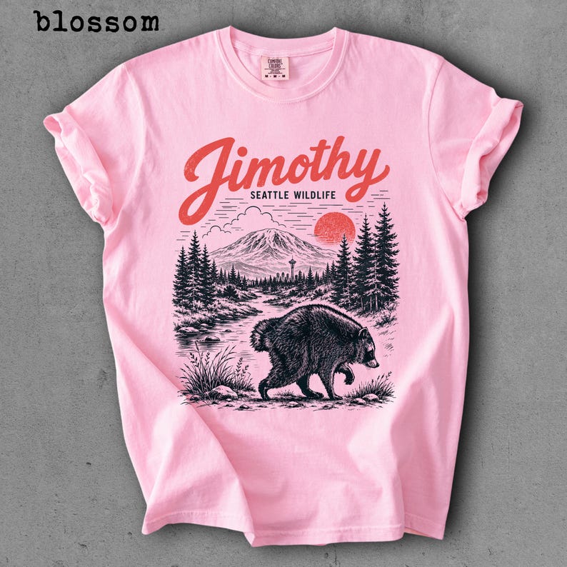 May include: A light pink t-shirt featuring a black and red graphic print. The design displays a bear walking near a river, pine trees, a mountain, a red sun, and the text "Jimothy SEATTLE WILDLIFE". The word "blossom" is printed in the top left corner.