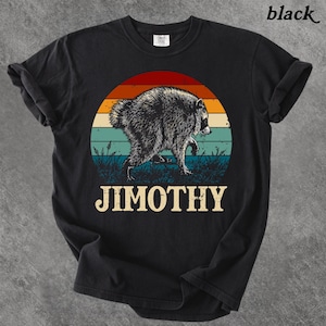 May include: A black t-shirt featuring a graphic of a raccoon walking through grass in front of a sunset with horizontal stripes in red, orange, yellow, and teal. The word JIMOTHY is printed in large, bold, cream-colored capital letters below the graphic.