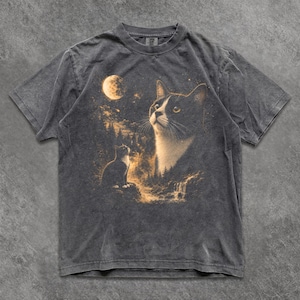 May include: A charcoal grey short-sleeve t-shirt featuring a graphic print of a black and white cat looking up at a full moon. The design includes a smaller cat silhouette, a forest landscape, and a waterfall in warm, golden-yellow tones.
