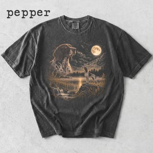 May include: A charcoal gray t-shirt with the word pepper printed in the top left corner. The center graphic features a spotted dog looking toward a full moon over a lake, with a second smaller dog standing on a rocky shore near the water.
