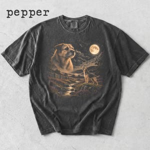 May include: A charcoal grey short-sleeve t-shirt with a crew neckline. The front features a graphic print of a dog looking at a full moon over a landscape with a stream and a second smaller dog. The word pepper is printed in black above the shirt.