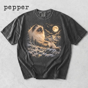 May include: A charcoal gray t-shirt featuring the text pepper in the upper left corner. The center graphic depicts a large, fluffy cat head in the sky above a snowy mountain landscape with a smaller cat sitting on a rock near a waterfall under a full moon.