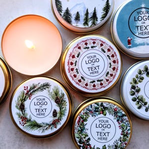 Puede incluir: Ocho latas redondas de velas con varios diseños de temática invernal. Cada lata presenta un fondo blanco con un diseño diferente, que incluye un bosque nevado, una corona de vegetación y un camión rojo. El texto "YOUR LOGO OR TEXT HERE" está impreso en cada lata.