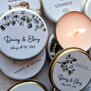 Può includere: Una collezione di piccole lattine rotonde per candele bianche con coperchi dorati. Le lattine sono decorate con un disegno ad acquerello di foglie verdi e il testo "Daisy & Ellery" e "August 03, 2023". Alcune delle lattine hanno una candela accesa all'interno.