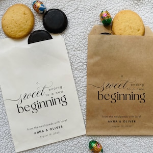 Op de afbeelding: Twee papieren traktatiezakjes, één crème en één bruin, met de tekst "a sweet ending to a new beginning". Elk zakje bevat een koekje en een chocoladetraktatie. Het crèmekleurige zakje heeft een donker chocoladekoekje.