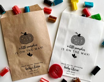 May include: Two brown paper favor bags with a pumpkin design and the text "a little pumpkin is on the way!"  The bags are filled with candy and have a thank you message for showering baby Olivia with love.