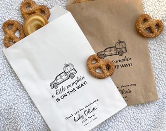May include: Two white paper bags with black text and illustrations. The text on the bags reads "a little pumpkin is on the way!" and "thank you for showering baby Olivia with love! August 19, 2025". The illustration is of a pickup truck with a pumpkin in the bed.