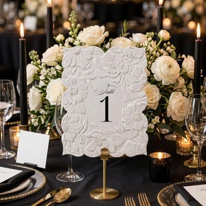 May include: A rectangular white table number sign featuring an embossed floral border and the text Table 1 in black script and serif font. The sign is mounted on a gold-toned metal stand with a circular base, positioned on a black tablecloth.
