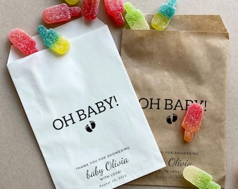 May include: Two white favour bags with the text "OH BABY!" printed on them. The bags are filled with colourful candy shaped like popsicles. The text "Thank you for showering baby Olivia with love! August 19, 2023" is printed on one bag. The text "U for showering Olivia with love! August 19, 2023" is printed on the other bag.