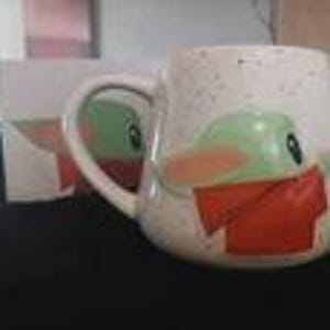 Puede incluir: Una taza de cer&aacute;mica blanca con acabado moteado que presenta el gr&aacute;fico de un personaje verde con ojos y orejas grandes que lleva una bufanda roja. La taza tiene un asa curva y una caja a juego visible en el fondo de la imagen.