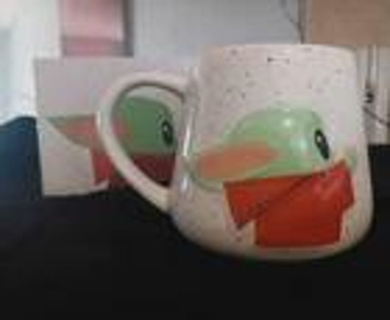 Puede incluir: Una taza de cer&aacute;mica blanca con acabado moteado que presenta el gr&aacute;fico de un personaje verde con ojos y orejas grandes que lleva una bufanda roja. La taza tiene un asa curva y una caja a juego visible en el fondo de la imagen.
