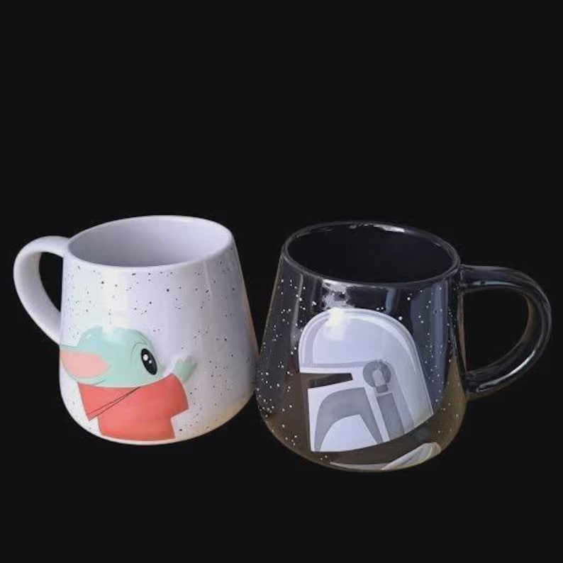 Puede incluir: Dos tazas de cer&aacute;mica con cuerpos redondeados y asas laterales. Una taza es blanca con motas negras y un gr&aacute;fico de un personaje verde con t&uacute;nica beige. La otra taza es negra con motas blancas y un gr&aacute;fico de un casco plateado con visor en T.