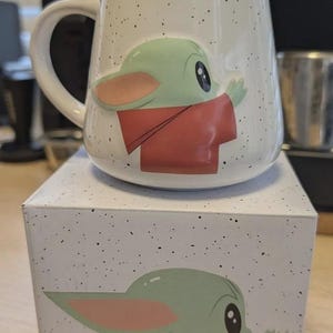 Puede incluir: Una taza de cer&aacute;mica blanca con acabado moteado que presenta un gr&aacute;fico en relieve de un personaje verde de orejas grandes con una t&uacute;nica roja. La taza se apoya sobre una caja cuadrada a juego que muestra la misma ilustraci&oacute;n del personaje.