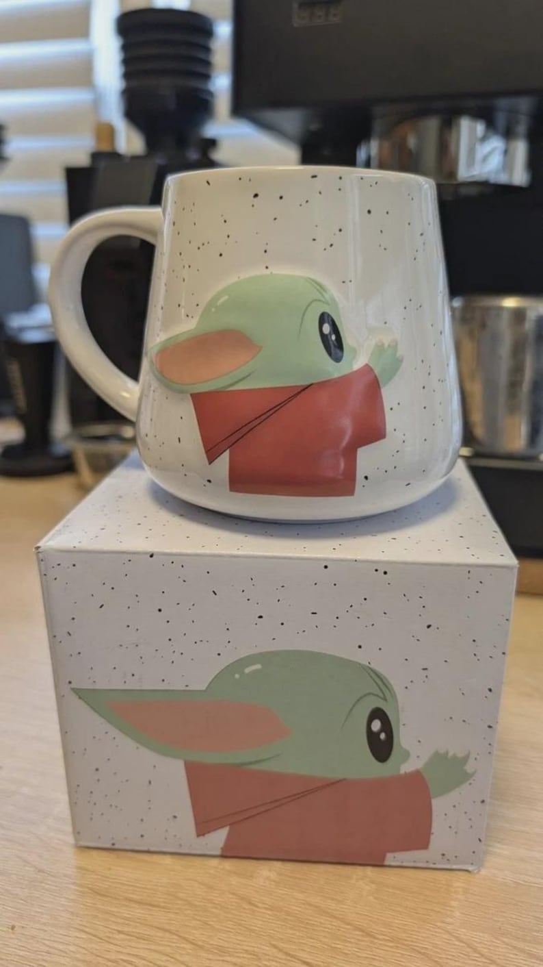 Puede incluir: Una taza de cer&aacute;mica blanca con acabado moteado que presenta un gr&aacute;fico en relieve de un personaje verde de orejas grandes con una t&uacute;nica roja. La taza se apoya sobre una caja cuadrada a juego que muestra la misma ilustraci&oacute;n del personaje.