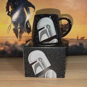 Puede incluir: Una taza de cer&aacute;mica negra con un gr&aacute;fico de casco estilizado en blanco y gris sobre un fondo estrellado. La taza se asienta sobre una caja de cart&oacute;n cuadrada a juego que muestra el mismo dise&ntilde;o de casco y patr&oacute;n de estrellas.