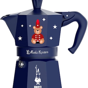 Puede incluir: Una cafetera Moka Express de color azul oscuro con un gráfico de un oso de peluche marrón vestido de soldado de juguete, rodeado de estrellas blancas. El cuerpo incluye el texto Moka Express y el logotipo de Bialetti con una válvula.