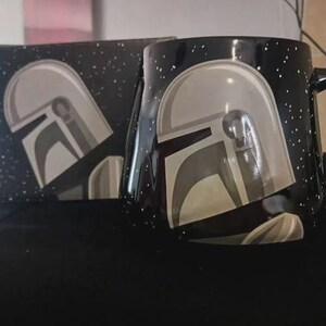 Puede incluir: Una taza de cer&aacute;mica negra con un patr&oacute;n de estrellas blancas moteadas y un gr&aacute;fico de un casco plateado. La taza est&aacute; colocada junto a una caja negra a juego que tambi&eacute;n muestra el patr&oacute;n de estrellas blancas y el mismo dise&ntilde;o de casco.