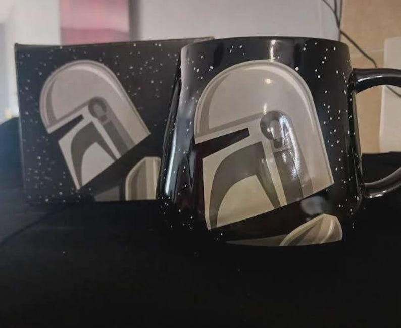 Puede incluir: Una taza de cer&aacute;mica negra con un patr&oacute;n de estrellas blancas moteadas y un gr&aacute;fico de un casco plateado. La taza est&aacute; colocada junto a una caja negra a juego que tambi&eacute;n muestra el patr&oacute;n de estrellas blancas y el mismo dise&ntilde;o de casco.