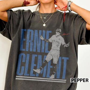 May include: Dark grey t-shirt with a graphic of baseball player Ernie Clement in a Blue Jays uniform. The name "ERNIE CLEMENT" is printed in blue block letters above the image of the player. The word "PEPPER" is printed in white in the bottom right corner.