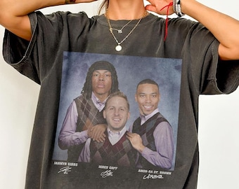 May include: Dark grey t-shirt featuring a group portrait of three men in argyle vests and collared shirts. The names "Jahmyr Gibbs", "Jared Goff", and "Amon-Ra St. Brown" are printed below the image, along with their signatures.