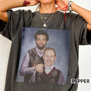 Op de afbeelding: Donkergrijs t-shirt met een retro portret van twee mannen in argyle vesten en overhemden met kraag. Het woord "PEPPER" is in het wit gedrukt in de rechter benedenhoek van het t-shirt.