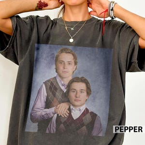 May include: Dark grey t-shirt with a vintage portrait of two young men in argyle vests and collared shirts. The word "PEPPER" is printed in white on the bottom right corner of the shirt.