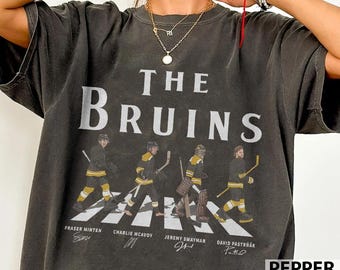 May include: Dark gray t-shirt featuring the text "THE BRUINS" above an image of four hockey players crossing a crosswalk. The players are wearing black and gold uniforms. Player names are printed below the image.