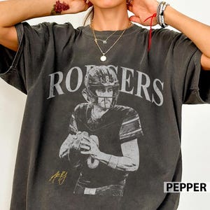 May include: Dark grey t-shirt with a grayscale image of a football player, the name "ROGERS" above. The player wears a helmet and holds a football. The shirt has the word "PEPPER" printed on the bottom right.