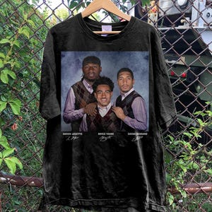 May include: Black t-shirt featuring a portrait of three people wearing argyle vests and collared shirts. The names Xavier Legette, Bryce Young, and Chuba Hubbard are printed below the image. The shirt is hanging on a wooden hanger.