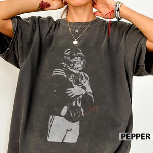 May include: Dark grey t-shirt featuring a grayscale image of a football player in a helmet and uniform. The player is holding a football. The shirt has the word "PEPPER" printed in white on the bottom right.