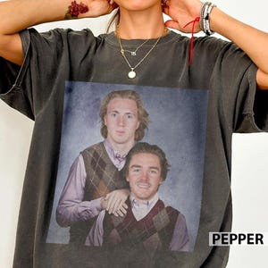 May include: Dark grey t-shirt with a vintage portrait of two men in argyle vests and collared shirts. The word "PEPPER" is printed in white on the bottom right corner of the shirt. The t-shirt is a casual garment.