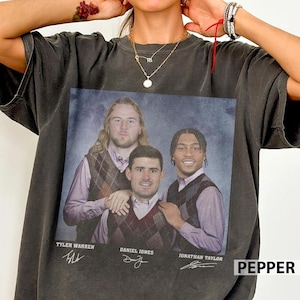 Puede incluir: Camiseta gris oscuro con una foto de grupo de tres hombres con chalecos de rombos y camisas con cuello. Los nombres Tyler Warren, Daniel Jones y Jonathan Taylor están impresos debajo, junto con sus firmas. La palabra "PEPPER" está impresa en blanco.