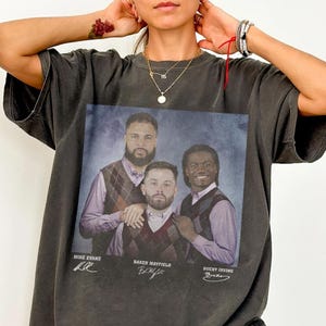 Puede incluir: Camiseta gris oscuro con un retrato de estilo vintage de tres hombres con chalecos de rombos y camisas con cuello. Los nombres "Mike Evans", "Baker Mayfield" y "Bucky Irving" están impresos debajo de la imagen en una fuente manuscrita.