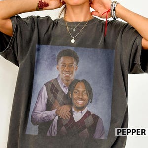 May include: Dark grey t-shirt with a portrait of two smiling young men in argyle vests over collared shirts. The word "PEPPER" is printed in white on the bottom right. The t-shirt has a vintage, worn appearance.