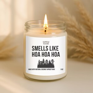 May include: A lit candle in a clear glass jar with a white label. The label reads "SMELLS LIKE HOA HOA HOA" with a forest graphic. The candle is made with natural coconut apricot wax and is 9 oz.