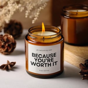 Könnte beinhalten: Eine brennende Sojawachskerze in einem klaren Glas mit braunem Farbton. Die Kerze hat ein weißes Etikett mit dem Text "BECAUSE YOU'RE WORTH IT" und "4 oz | 20 hrs".