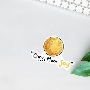 May include: A sticker featuring a yellow cartoon moon with craters and a black outline. The sticker has the text "Copy, Moon joy" in black and yellow. The sticker is on a white surface.