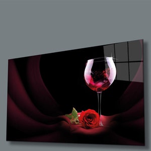 May include: A single red rose with green leaves lies on a dark red fabric next to a glass of red wine. The wine glass is clear and has a round bowl. The background is black.